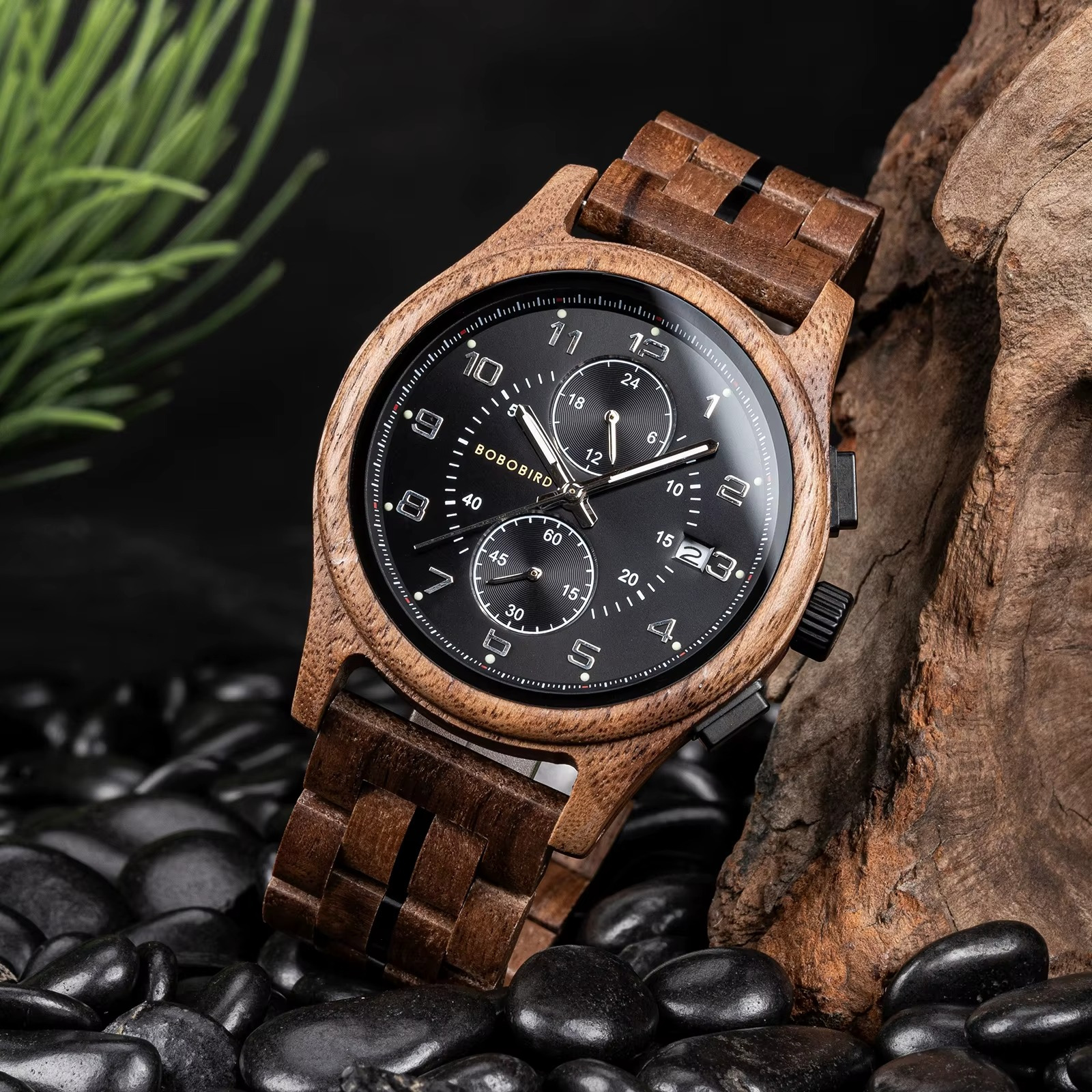 Montre en bois pour homme Yukon avec cadran noir chronographe et bracelet en noyer posée sur des pierres noires à côté d'un morceau de bois