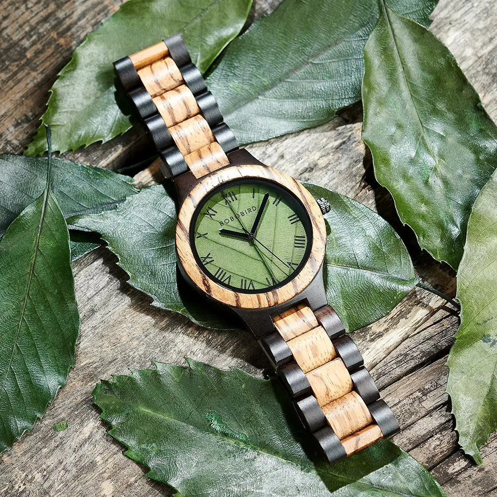 Montre en bois Sylva avec cadran vert feuille et chiffres romains, bracelet en bois clair et foncé, posée sur des feuilles vertes